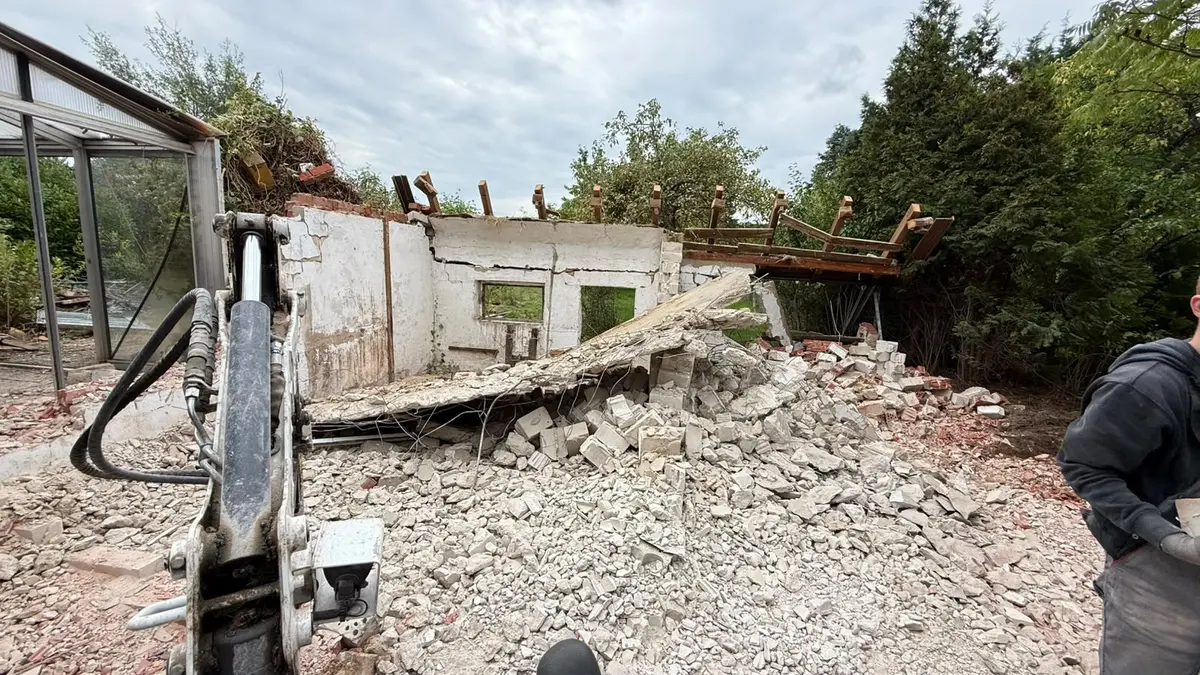 Baggerarm vor der letzten stehenden Wand, daneben ein Mitarbeiter